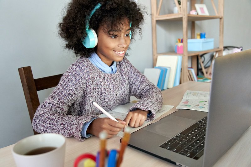 Cute happy african school kid girl wearing headphones virtual distance learning online watching remote digital class lesson looking at laptop computer studying at home sitting at desk, writing notes