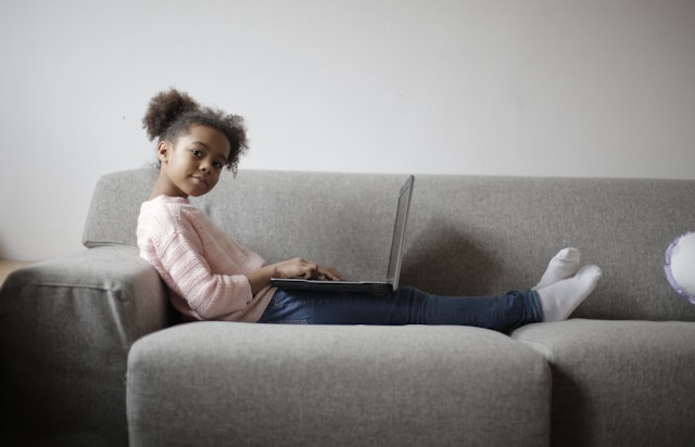 girl in pink long sleeve shirt and black pants sitting on gray couch with laptop